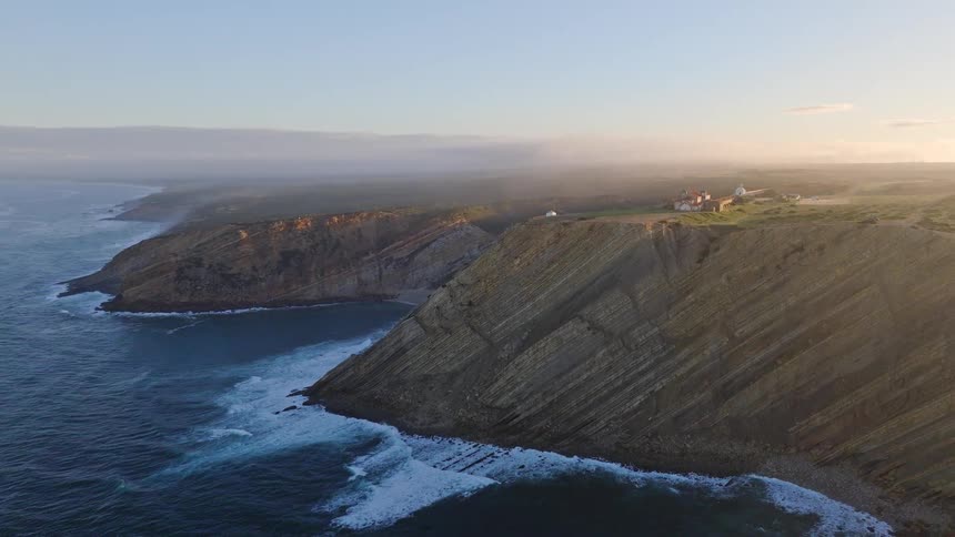 A stunning aerial view of a rugged, misty coastline at sunrise, revealing dramatic layered cliffs and a sanctuary on the clifftop