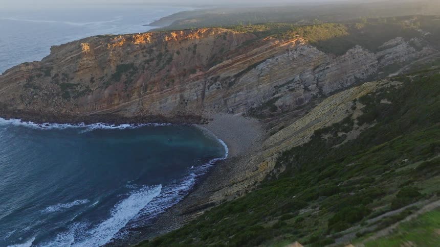 An aerial view pulls back from a secluded cove with crashing waves to reveal a small, white chapel on a dramatic cliffside at sunrise