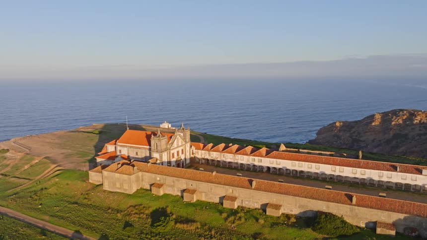 Cinematic aerial view of the historic Sanctuary of Our Lady of Cape Espichel on a grassy headland overlooking the Atlantic Ocean