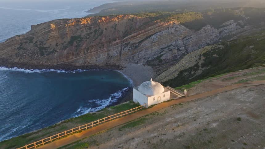 An awe-inspiring aerial view captures the historic Ermida da Memória chapel sitting majestically on the cliffs of Cabo Espichel, Portugal, during a golden sunset