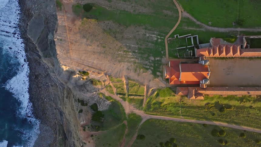 A stunning top-down aerial view of the Santuário de Nossa Senhora do Cabo Espichel perched on a dramatic cliffside during a beautiful golden hour sunset