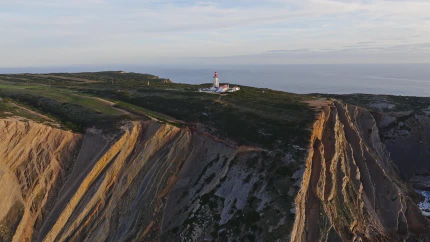 A majestic aerial shot ascends over stunning coastal cliffs, revealing a solitary lighthouse overlooking the vast ocean during the golden hour