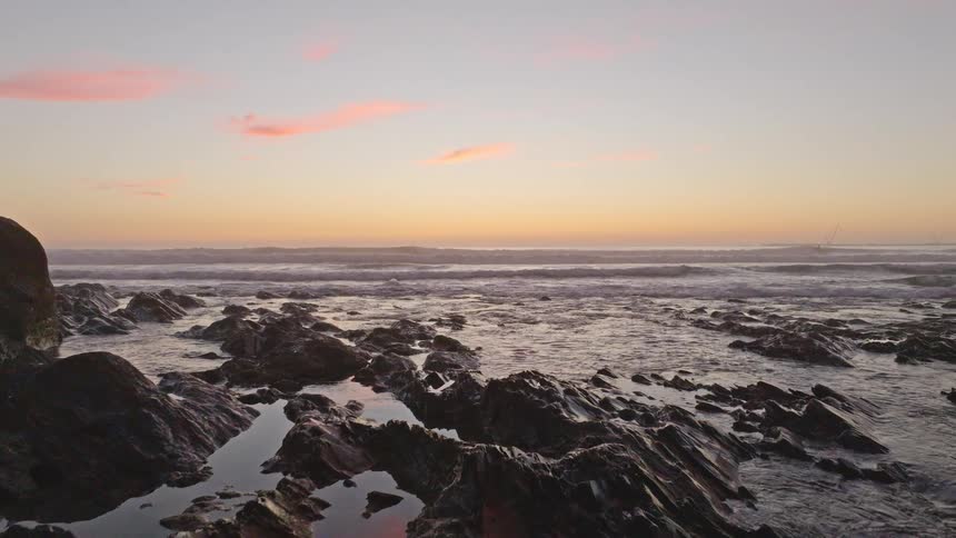 A slow pan reveals a serene, rocky coastline with gentle ocean waves under a beautiful sunset sky with hints of pink