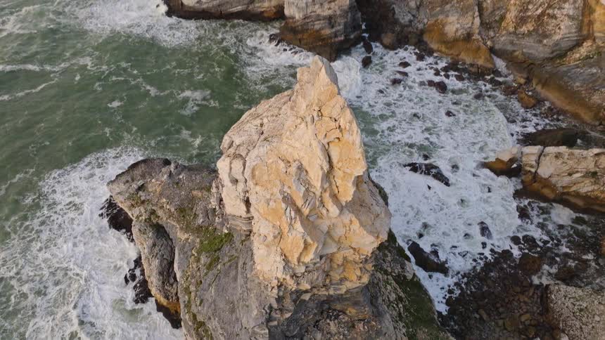 Aerial top-down view pulls back to reveal a dramatic, rugged rock formation amidst crashing ocean waves on a stunning coastline