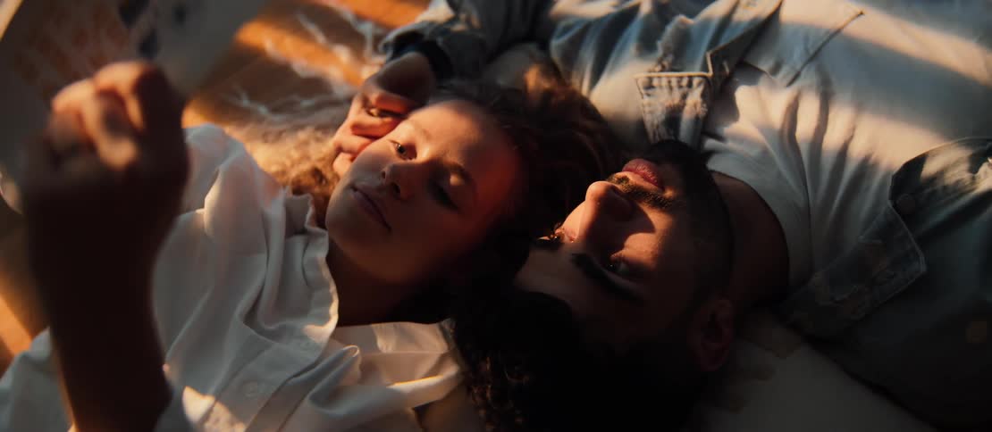A young couple lays close together on a bed in the warm afternoon sun while looking at an old photograph