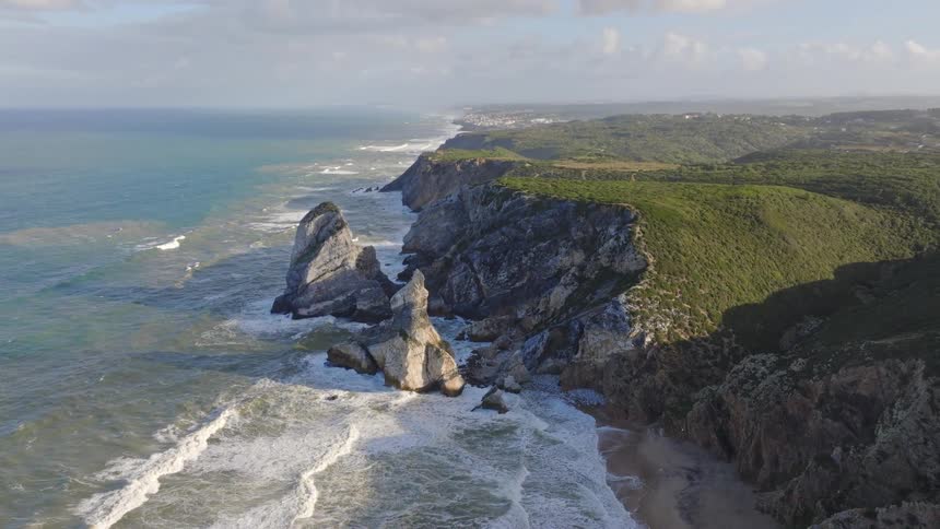 Slowly pulling back aerial shot captures the majestic beauty of Portugal's rocky coastline with large sea stacks and green cliffs