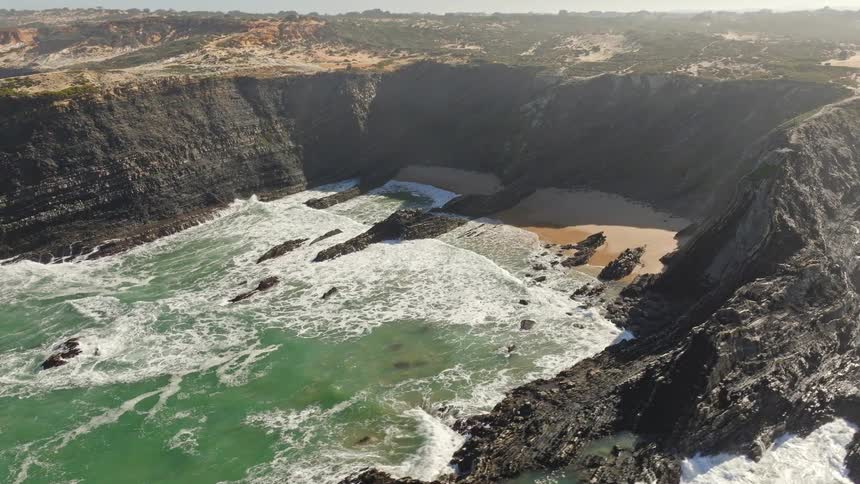 Flying backward over a stunning coastal landscape reveals a secluded cove with a sandy beach and turquoise waves crashing against dark, dramatic cliffs