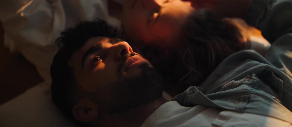 A young man contemplates peacefully while lying in bed next to his partner, their faces illuminated by warm morning sunlight