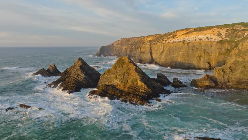 An aerial view captures the dramatic rocky coastline as powerful waves crash against the sea stacks during a beautiful golden hour