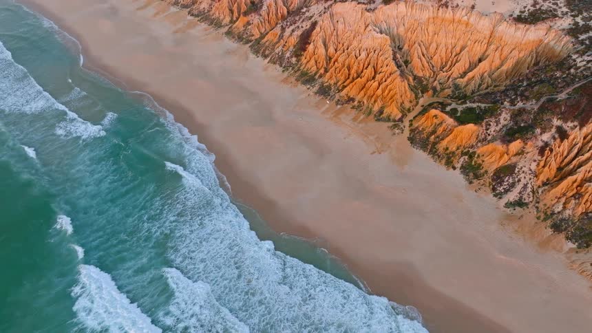 A stunning aerial view starts with a top-down shot of turquoise waves and a sandy beach beside vibrant orange eroded cliffs, then pulls back to reveal the vast coastline