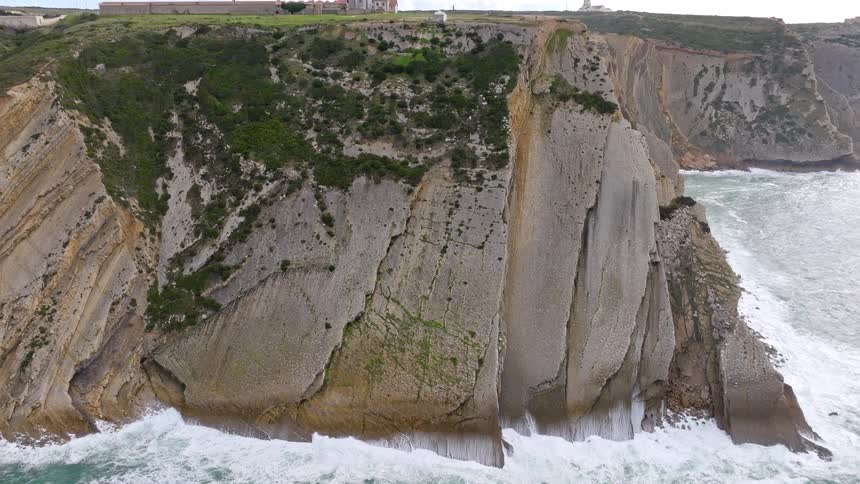 A dramatic aerial reveal shot pulls back from a massive cliff face to show the Cabo Espichel lighthouse and sanctuary on the rugged Portuguese coastline