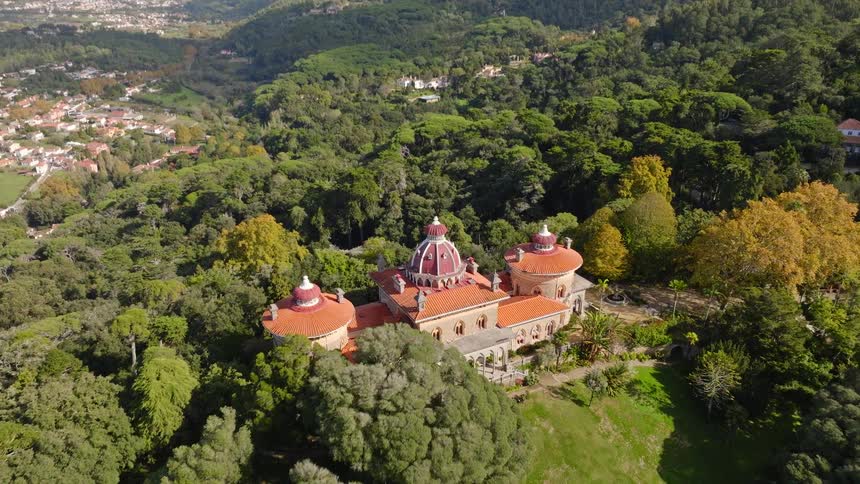 An aerial view slowly tilts down over the magnificent Monserrate Palace with its iconic red domes, nestled in the lush forests of Sintra, Portugal