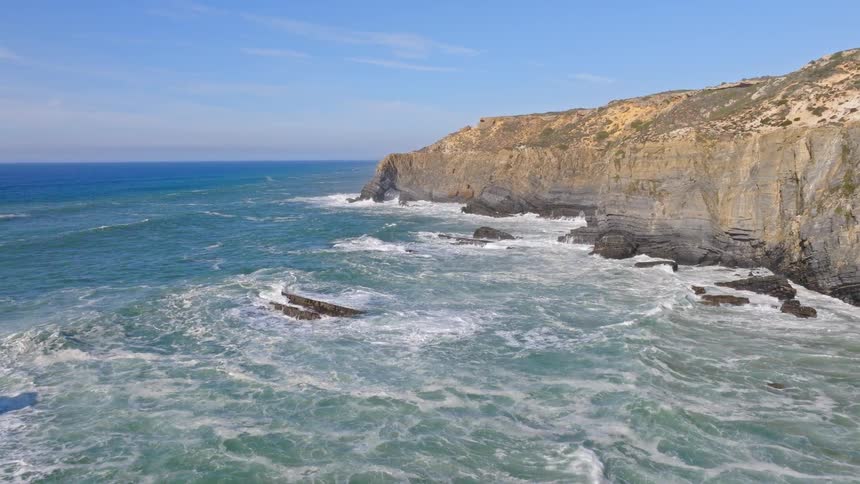 An epic aerial view flies over rugged sea stacks along a dramatic cliffside coastline with powerful ocean waves crashing below