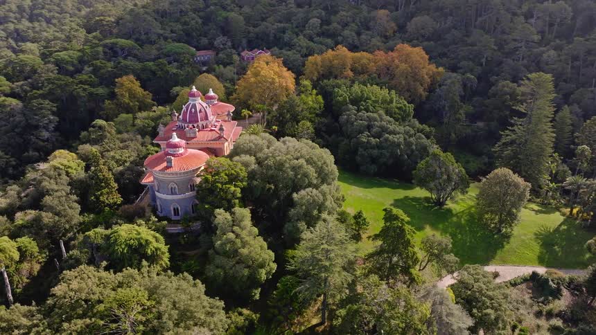 An ornate, exotic palace with distinctive red domes sits peacefully surrounded by a vast, lush forest and manicured parkland