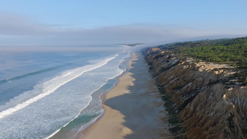 Aerial view pans over a foggy ocean with waves rolling onto a sandy beach alongside dramatic eroded cliffs topped with a green forest