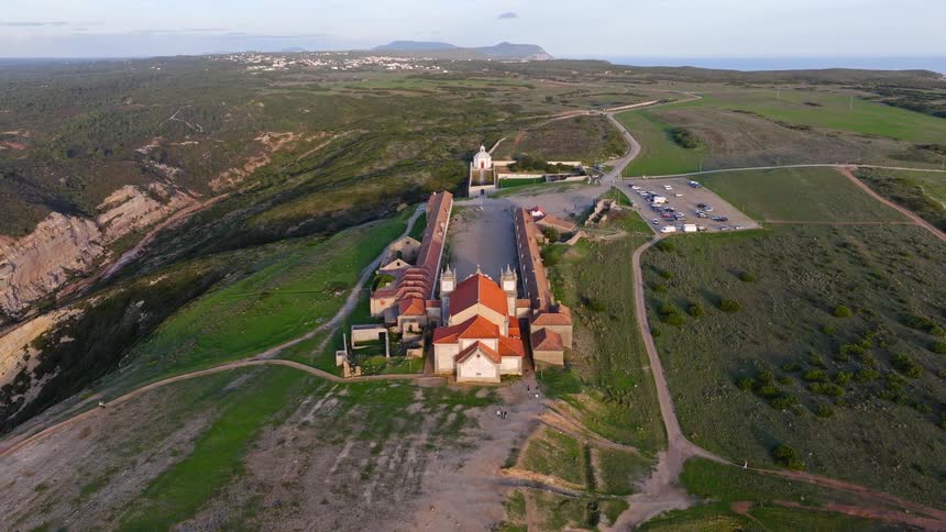 An aerial top-down shot slowly pulls back to reveal the magnificent Santuario de Nossa Senhora do Cabo Espichel monastery perched on a dramatic cliffside in Portugal