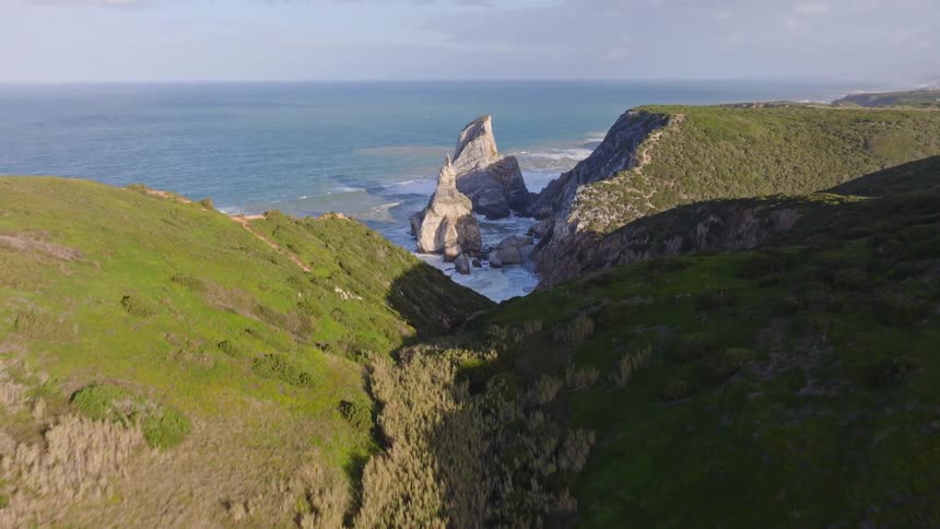 Aerial drone footage flies through a lush green valley revealing the dramatic Ursa Beach and its striking rock formations on the Portuguese coast