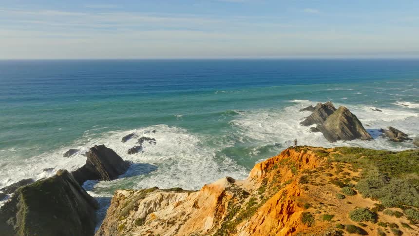 An epic aerial view pulls back to reveal a lone person standing on a rugged cliff overlooking the vast, churning ocean and dramatic coastline