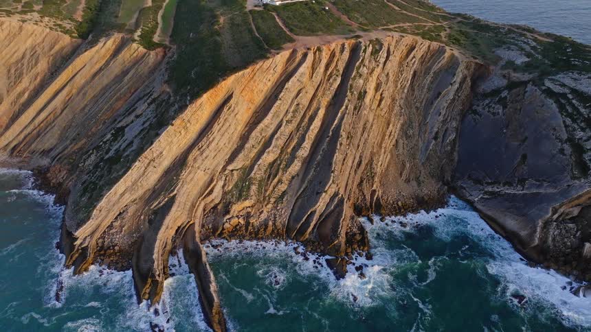 An aerial drone shot pulls back from the turbulent ocean waves crashing against unique layered cliffs to reveal a stunning lighthouse perched on the coast
