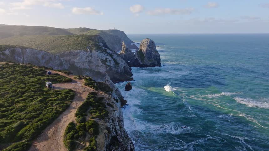 An epic aerial view slowly flies forward along a stunning green, rocky coastline as powerful ocean waves crash against the cliffs