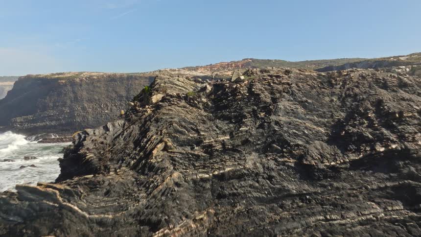An aerial drone shot flies over a rugged, rocky coastline revealing a lone person standing on the cliff's edge overlooking a secluded beach cove