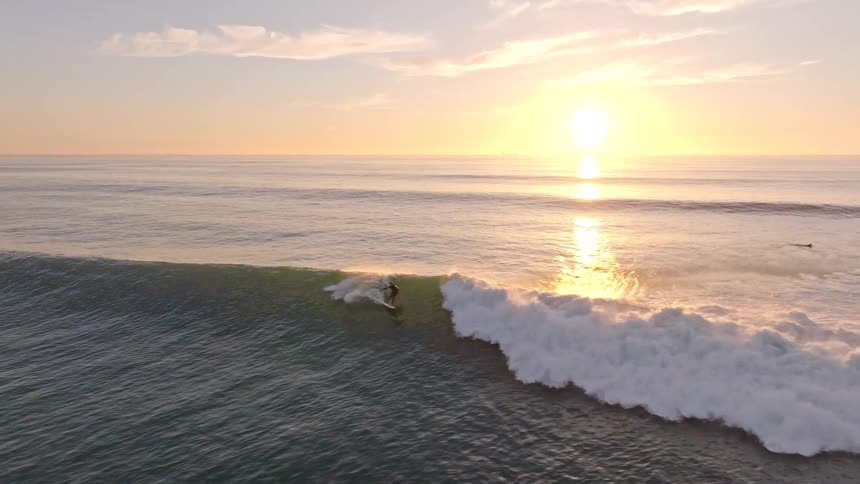 An aerial slow-motion shot follows a surfer gliding along a perfect ocean wave as the golden sun rises over the horizon