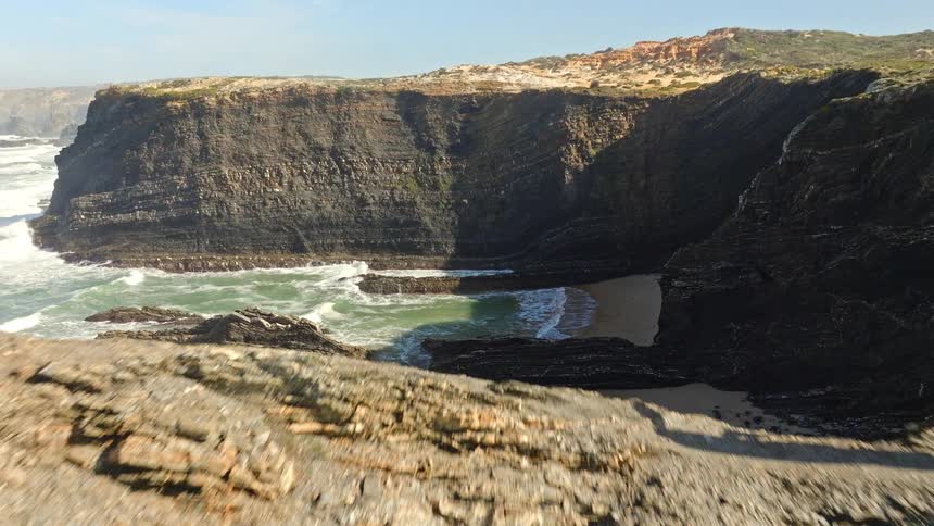 Aerial shot pulls back from a secluded beach to reveal a lone man standing on a dramatic, rocky cliff overlooking the ocean