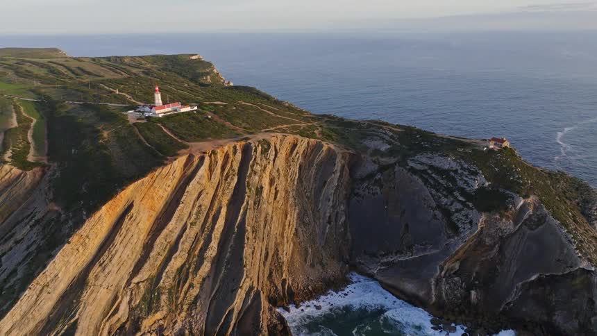 An aerial view reveals the majestic Cabo Espichel lighthouse perched atop dramatic, golden-lit cliffs as waves crash against the rocky shore