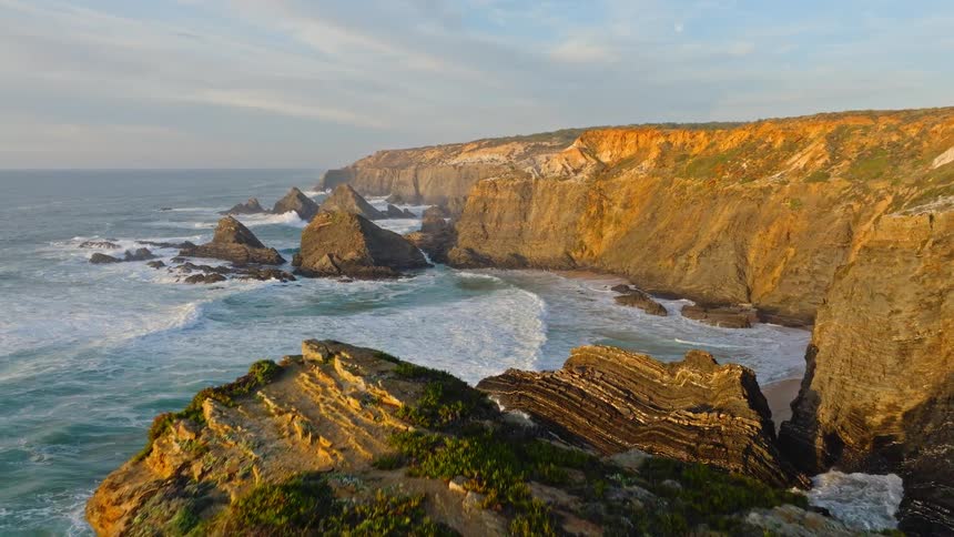 An aerial view rises over a rugged, rocky coastline where the ocean waves crash against the cliffs during a golden sunset