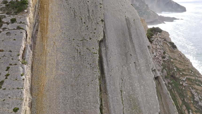 A detailed high-angle view captures the immense scale and texture of a stratified rock cliff face overlooking the ocean