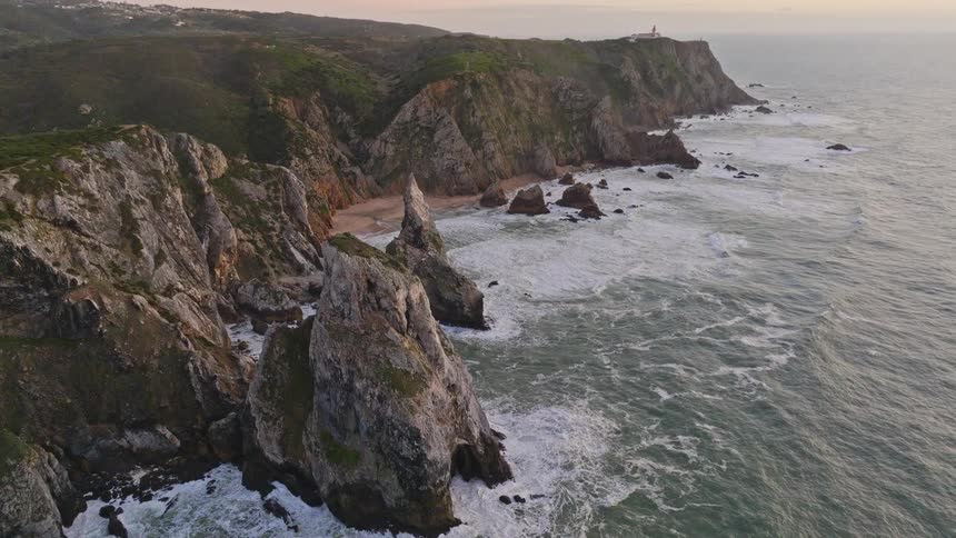 An epic aerial shot reveals a dramatic, rocky coastline with waves crashing against cliffs and a lighthouse visible in the distance at sunset