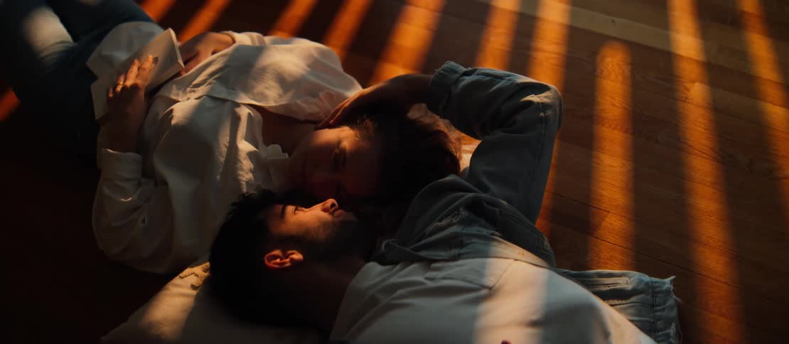 An affectionate couple lies together on the hardwood floor, bathed in the warm, striped light of the golden hour sun