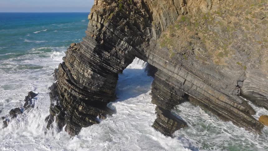 An incredible aerial shot pulls back from a massive natural rock arch as powerful ocean waves crash through it on a sunny day