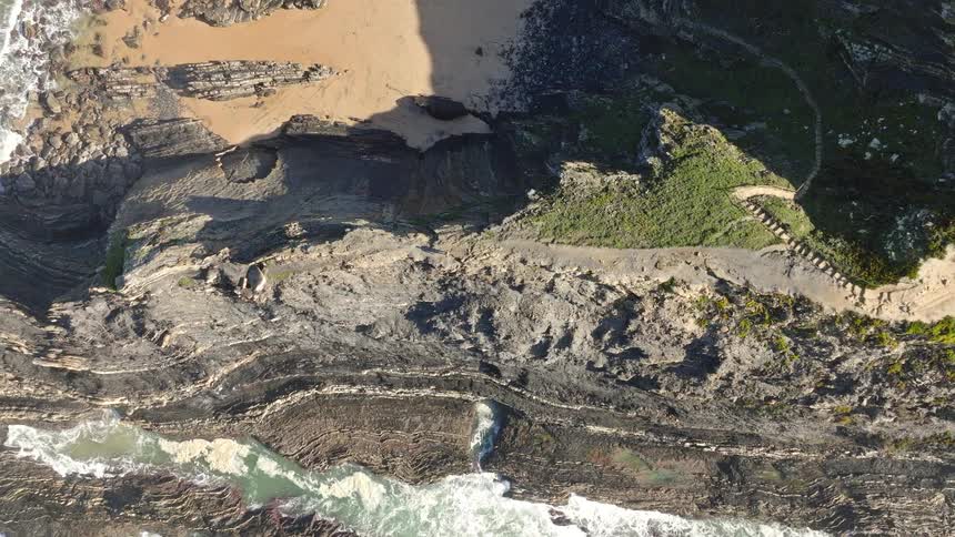 An aerial top-down view zooms out to reveal a lone hiker on a dramatic, rocky coastal path beside the crashing ocean waves