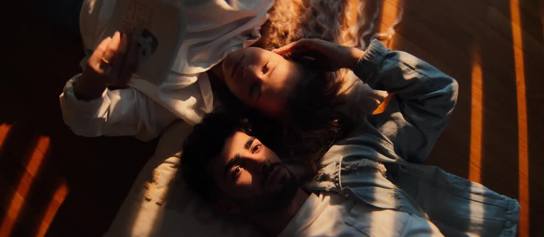 An intimate top-down shot of a young couple relaxing on the wooden floor, bathed in warm golden hour sunlight while the woman reads a book