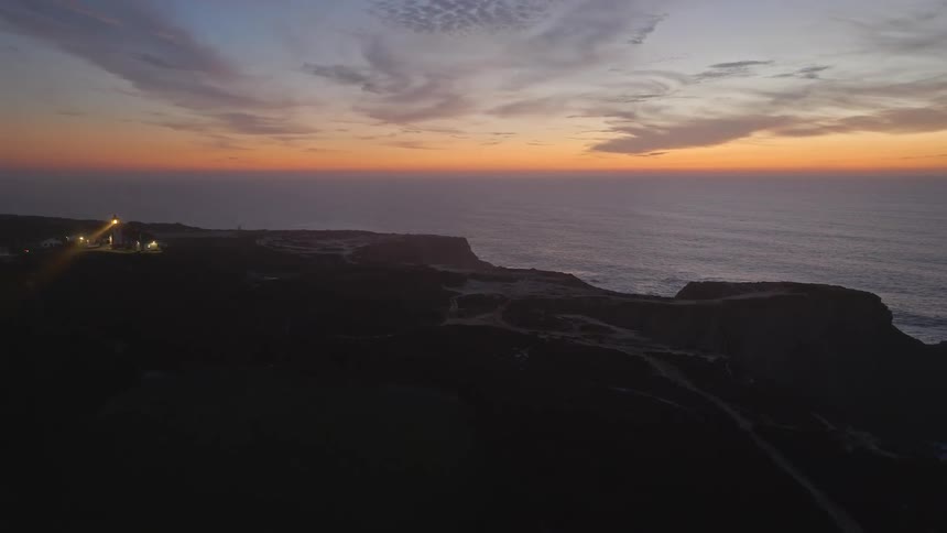 An aerial view shows a lighthouse turning on its beacon and shining light over the rugged coastline cliffs at sunset