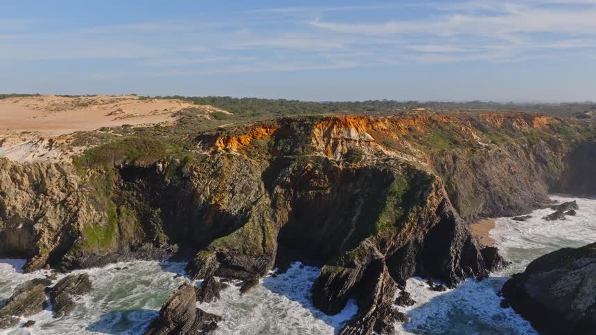 An aerial drone shot pulls back to reveal a beautiful and rugged coastline with dramatic cliffs, crashing waves, and flying birds