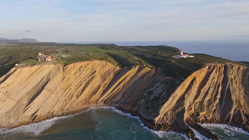 An aerial pan reveals the majestic layered cliffs, historic sanctuary, and lighthouse of Cabo Espichel, Portugal, bathed in the warm light of sunset