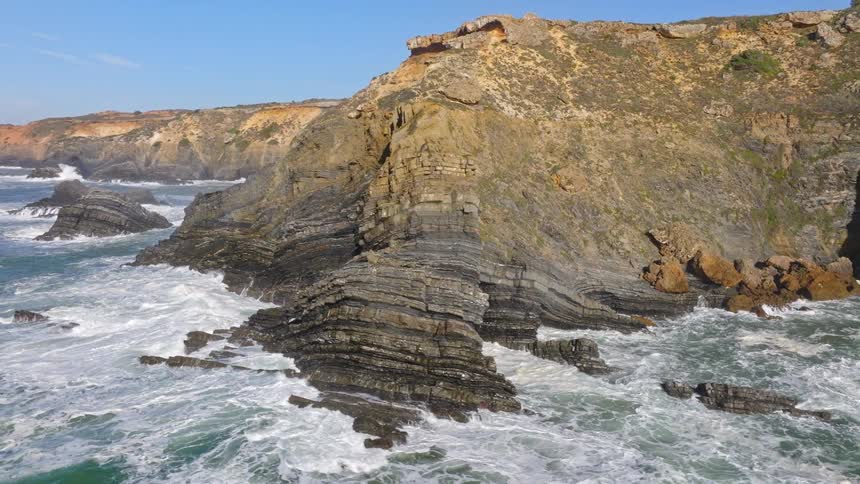 An aerial drone slowly pulls back from a magnificent sea arch, revealing the vast and rugged coastline as powerful ocean waves crash against the cliffs