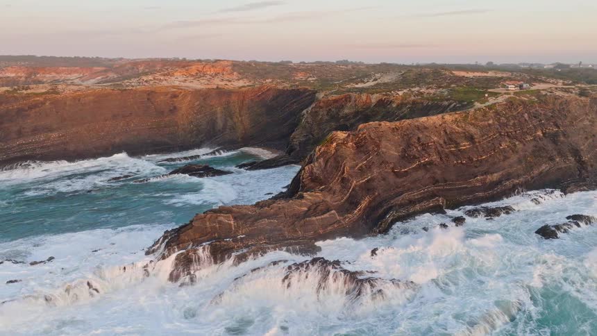 An aerial view reveals the rugged and dramatic coastline as powerful ocean waves crash against the rocky cliffs during a golden sunset