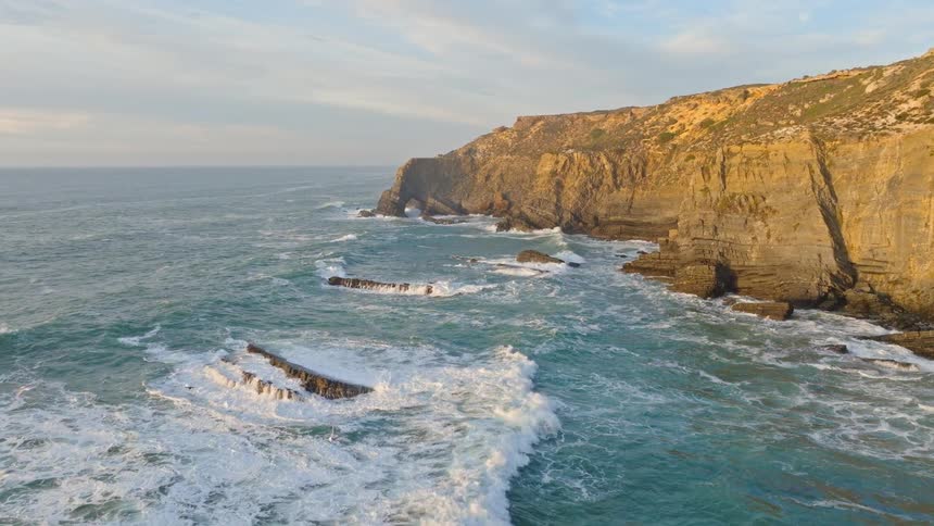 A majestic aerial flight over dramatic sea stacks reveals a rugged coastline with a natural arch as powerful ocean waves crash ashore during sunset