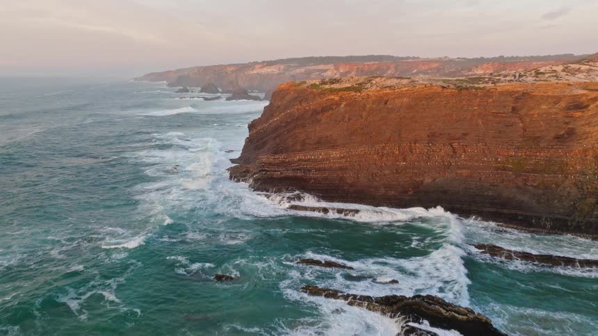 Dramatic aerial footage pulls back over rugged coastal cliffs as powerful ocean waves crash against the rocks at sunset