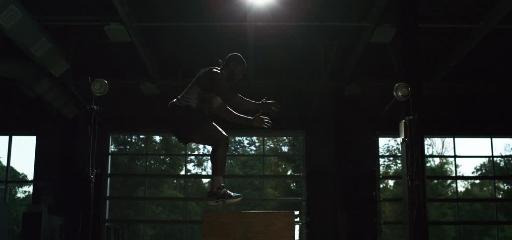 A muscular athlete performs a box jump in super slow motion in a dramatically lit gym