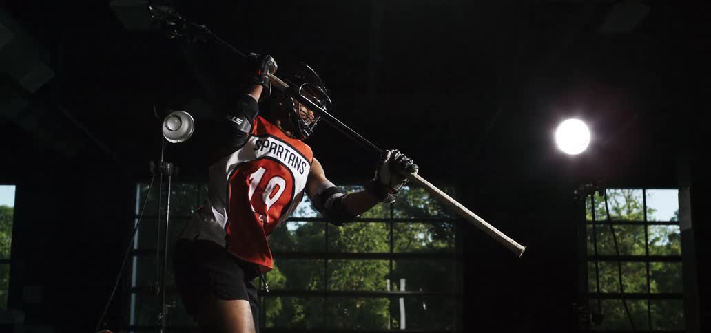 A woman in full lacrosse gear throws a ball in super slow motion, dramatically lit by a flash of light in a dark room