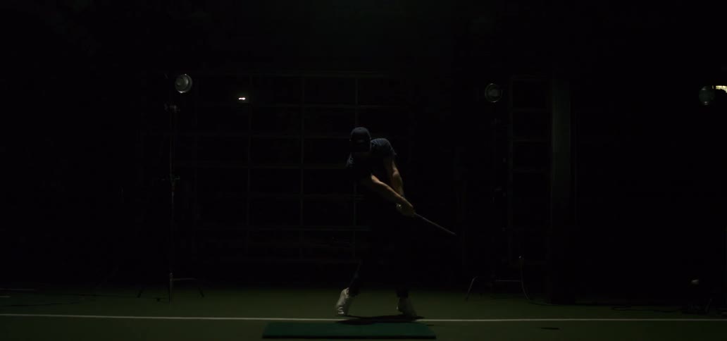 A golfer in a hat swings his club and hits a ball in slow motion inside a dramatically lit indoor simulator