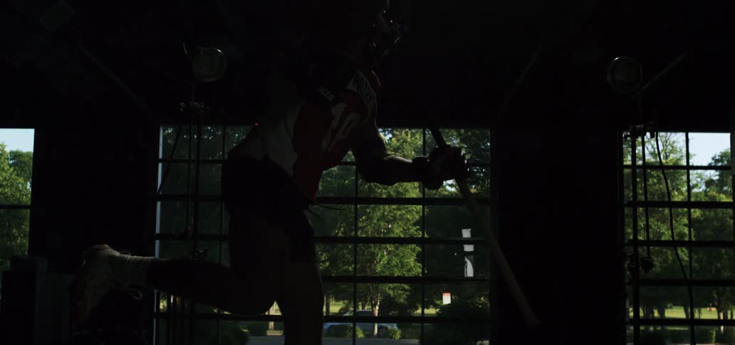 A lacrosse player is silhouetted against a large window before a bright light reveals him jumping and taking a powerful shot in dramatic super slow motion