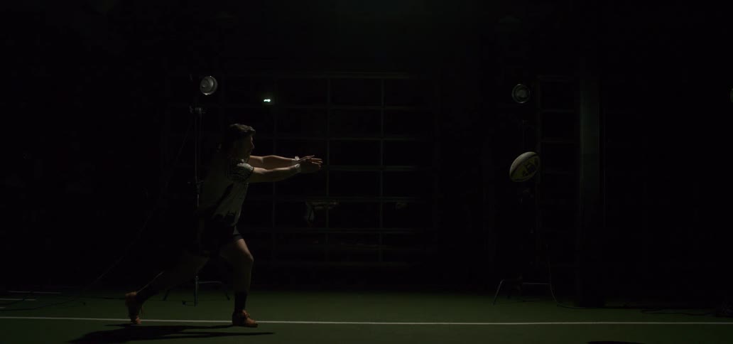 A male rugby player throws a pass in super slow motion, illuminated by flashing strobe lights in a dark indoor training facility