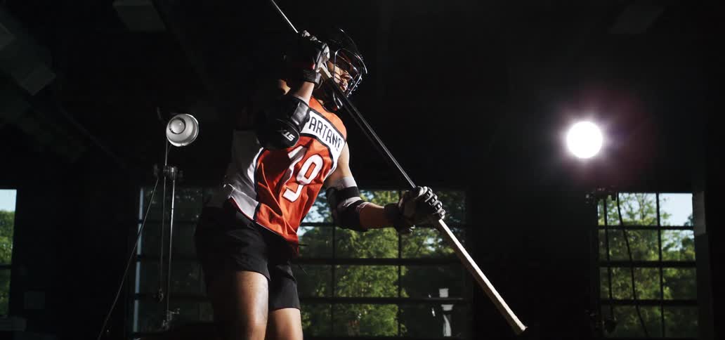 In a slow-motion sequence, a lacrosse player stands poised with his stick as dramatic lighting flashes in a dark studio