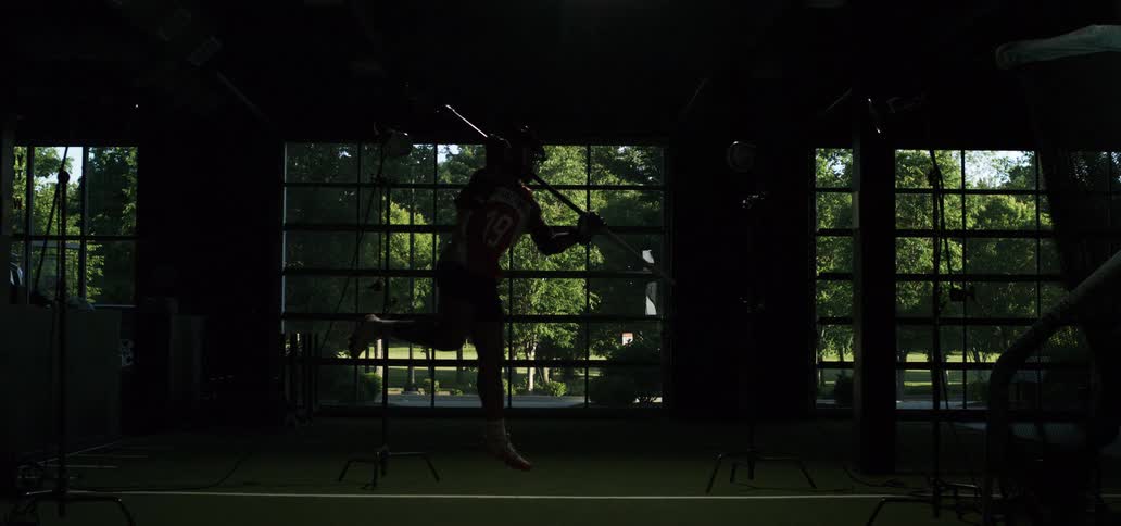 A lacrosse player jumps and runs in slow motion, dramatically revealed from silhouette by a bright flash of light