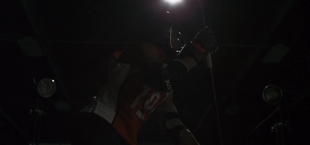A lacrosse player is dramatically lit by a spotlight while posing with his stick in slow motion